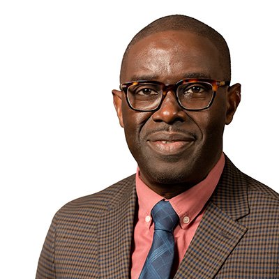A man with glasses and a patterned coat smiles confidently, wearing a pink shirt and blue tie against a white background.