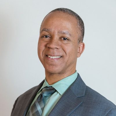 A smiling man dressed in a suit and tie, with short hair and a light blue shirt, stands against a neutral background.